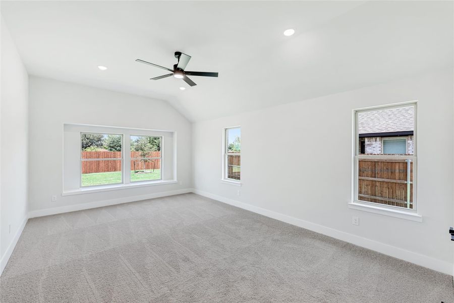 Empty bedroom with vaulted ceiling, ceiling fan, carpet flooring, and three windows overlooking backyard