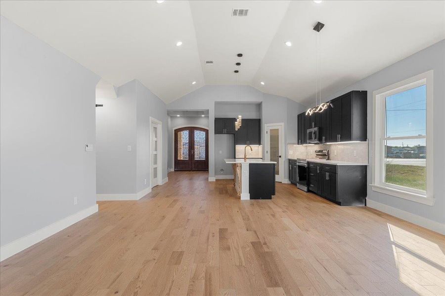 Kitchen with dark cabinetry, appliances with stainless steel finishes, an island with sink, open floor plan, and light wood finished floors Kitchen with dark cabinetry, appliances with stainless steel finishes, an island with sink, open floor plan, and light wood finished floors