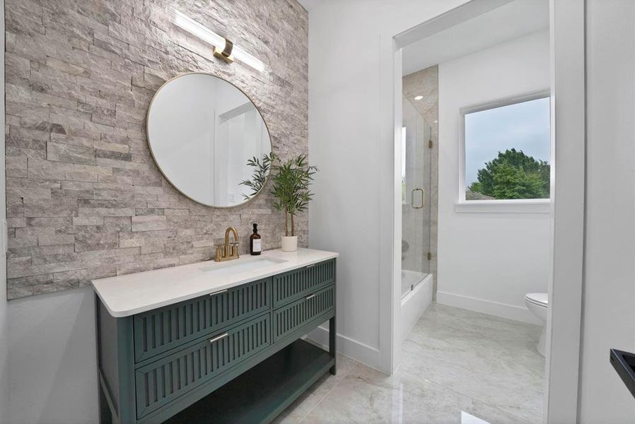 Contemporary bathroom featuring a stylish vanity with a white countertop and gold-toned fixtures, a circular mirror, and a textured stone accent wall