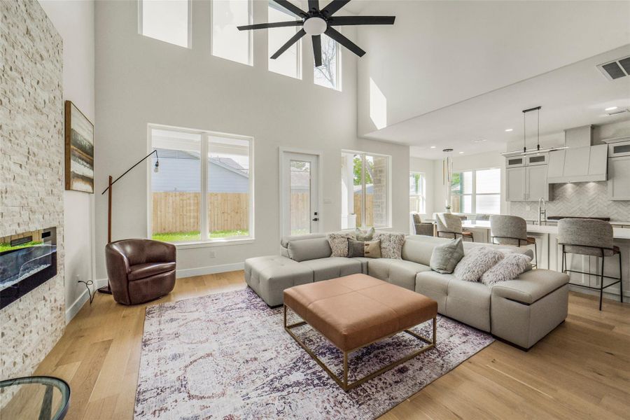 This spacious living area features high ceilings, abundant natural light, and a modern open layout. It includes a cozy sectional sofa, a stylish ottoman, and a sleek fireplace. The adjacent kitchen boasts contemporary finishes and a large island with seating, perfect for entertaining.