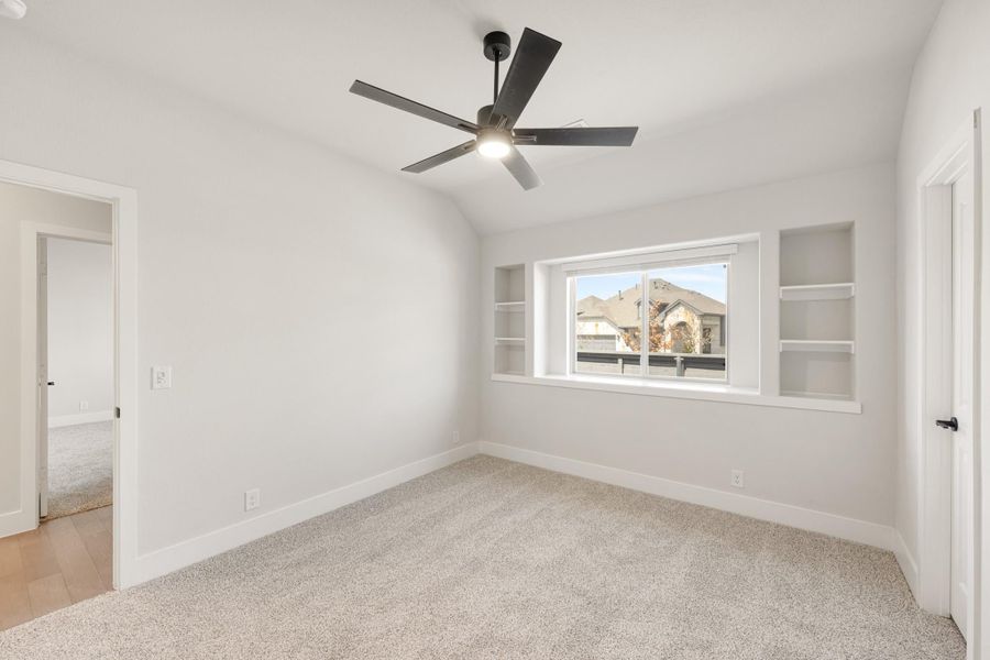 Empty bedroom with vaulted ceiling, ceiling fan, built-in shelves flanking a window, and carpet flooring