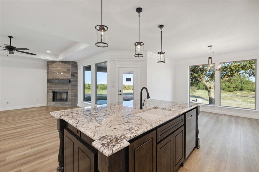 Kitchen with light wood-type flooring, an island with sink, a stone fireplace, decorative light fixtures, and sink