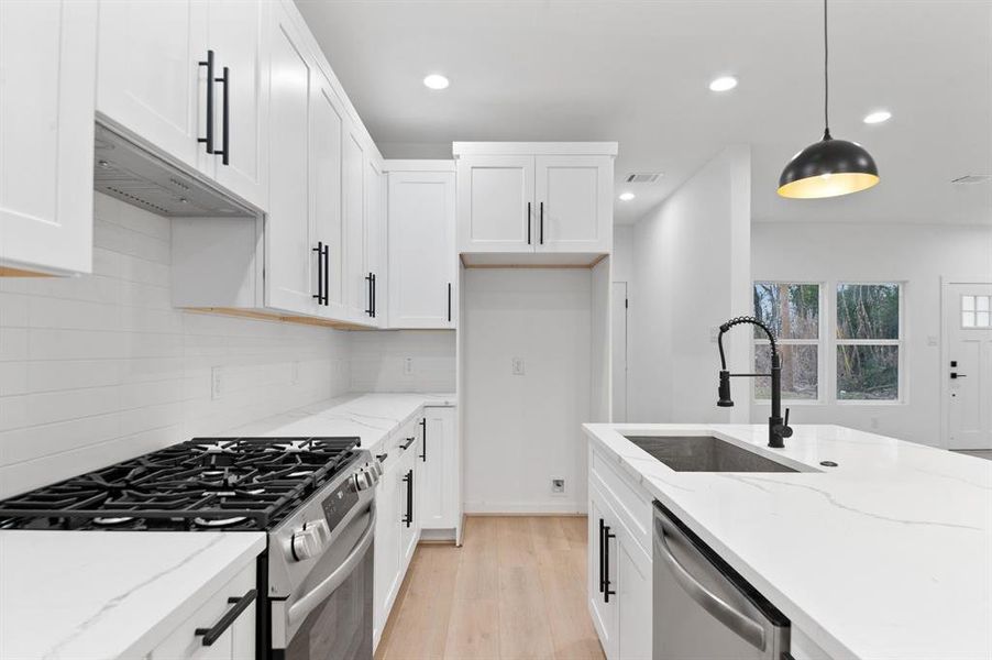 This modern kitchen features sleek white cabinetry, a spacious island with a dual sink, and stainless steel appliances, including a gas stove. The room is well-lit with recessed lighting and a stylish pendant light, complemented by large windows offering natural light.