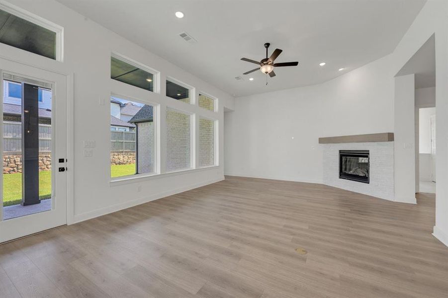 Spacious living area featuring light wood-finish flooring, a white brick fireplace with a wood mantle, and a ceiling fan