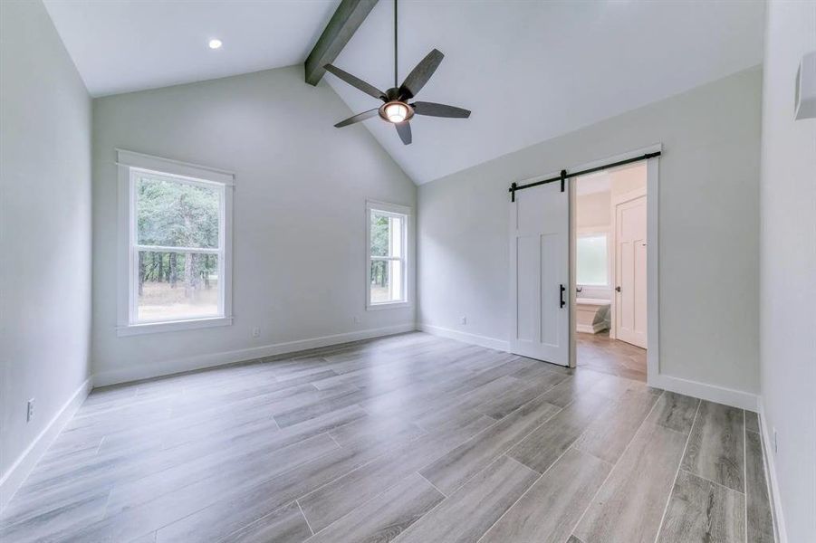 Empty room with lofted ceiling with beams, light hardwood / wood-style flooring, a barn door, and ceiling fan