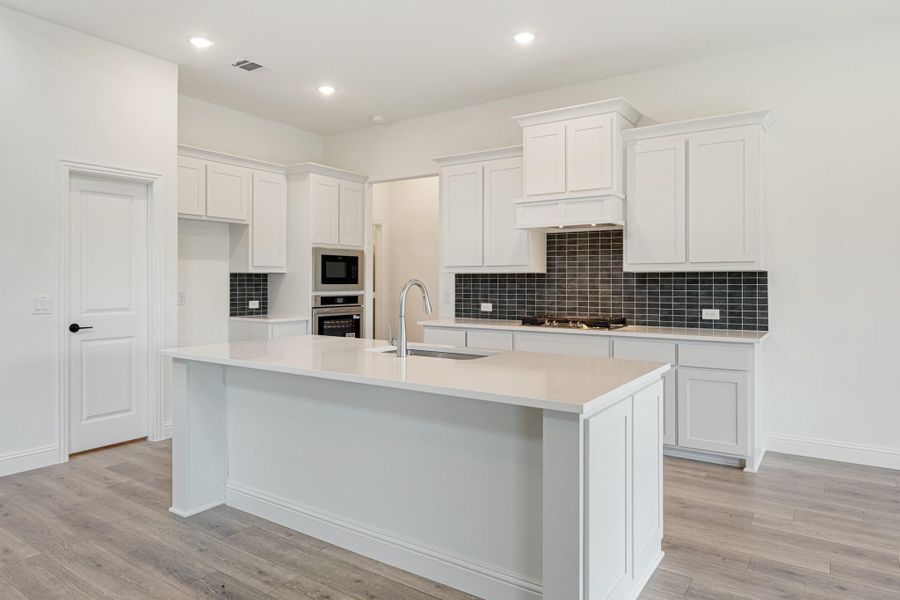 Kitchen with white cabinets, dark tile backsplash, center island with sink, and stainless steel appliances on light wood floors Kitchen with white cabinets, dark tile backsplash, center island with sink, and stainless steel appliances on light wood floors