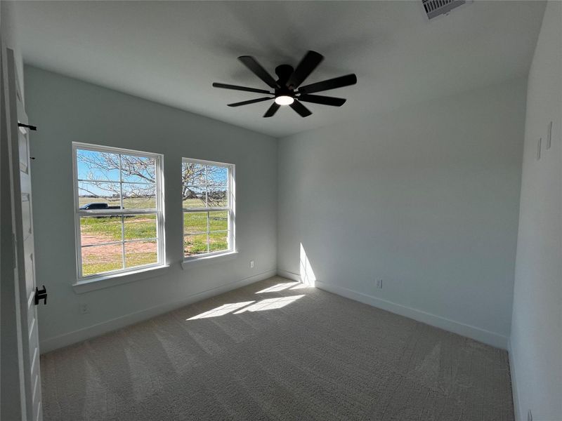 The first room to the left when walking in features a large window with views of a grassy landscape, allowing plenty of natural light. It has a neutral color palette, carpeted floor, and a modern ceiling fan, creating a bright and airy space.