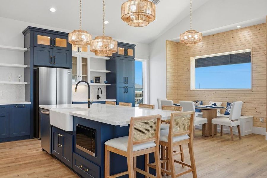 This photo features a modern kitchen with navy cabinets, a spacious island with a sink, and light wood flooring. There are elegant pendant lights and a cozy dining nook with a wooden accent wall, providing a warm and inviting atmosphere.