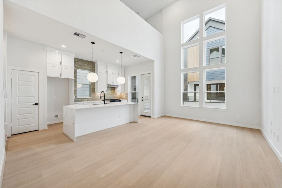 Open-concept living and dining area offers seamless flow and abundant natural light. Clean lines, neutral tones, and light wood floors create a bright, modern atmosphere that connects effortlessly to the kitchen for relaxed entertaining. Three stacked high windows and soaring 20-ft ceilings amplify the brightness and elevate the space’s airy, contemporary feel.