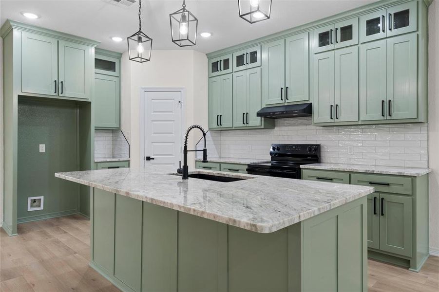 Kitchen with green cabinets, light stone counters, black range with electric cooktop, hanging light fixtures, and an island with sink
