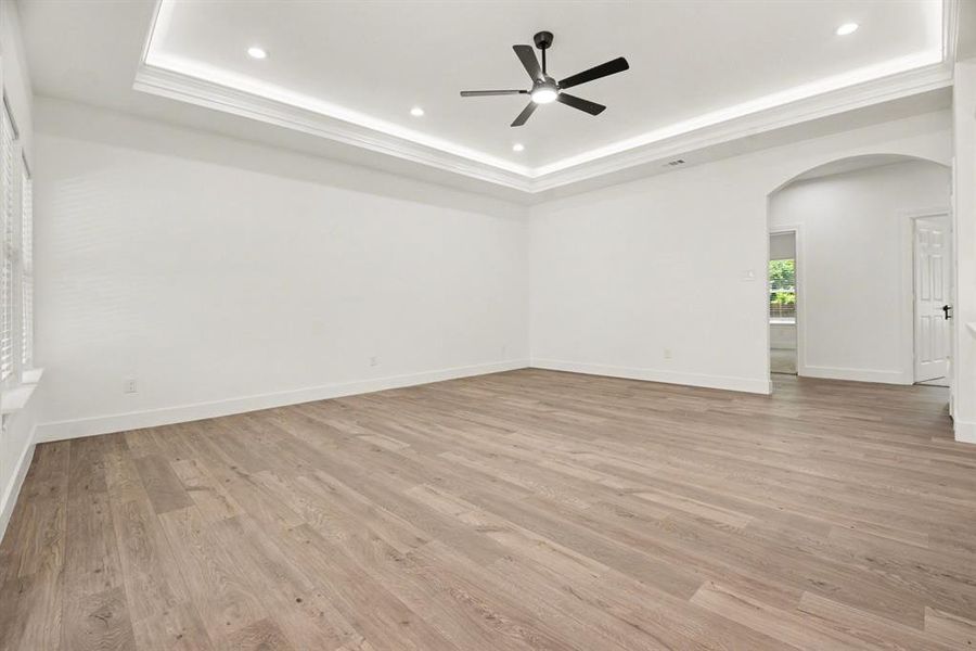 This expansive room features light-colored plank flooring, a tray ceiling with recessed lighting and a ceiling fan, and white walls