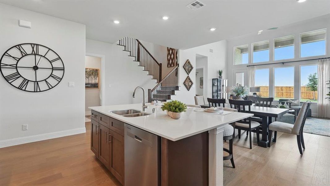 Open-concept living area featuring wood-finish flooring, a kitchen island with a stainless steel sink and integrated dishwasher, a formal dining space, and a two-story wall of windows