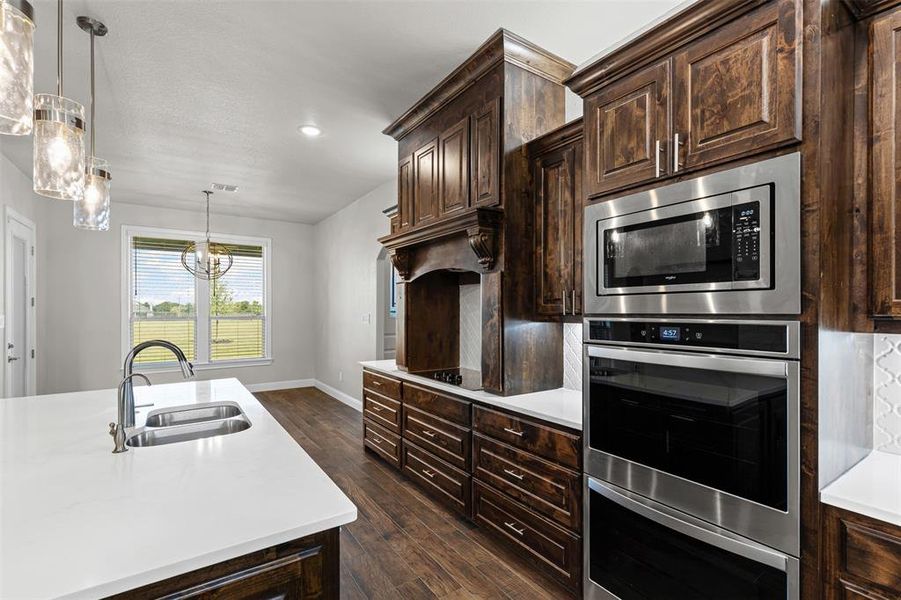 Kitchen featuring dark brown cabinetry, hanging light fixtures, stainless steel appliances, dark wood-style flooring, and light stone countertops Kitchen featuring dark brown cabinetry, hanging light fixtures, stainless steel appliances, dark wood-style flooring, and light stone countertops