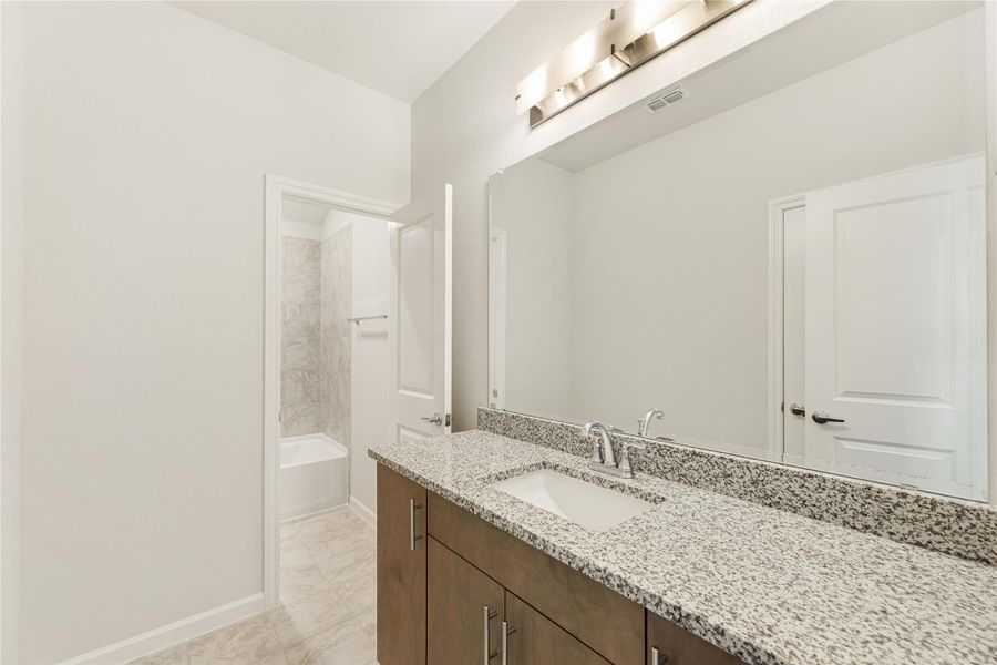 Bathroom with granite countertop, undermount sink, dark wood vanity, large mirror, and tiled shower-tub combo in background.