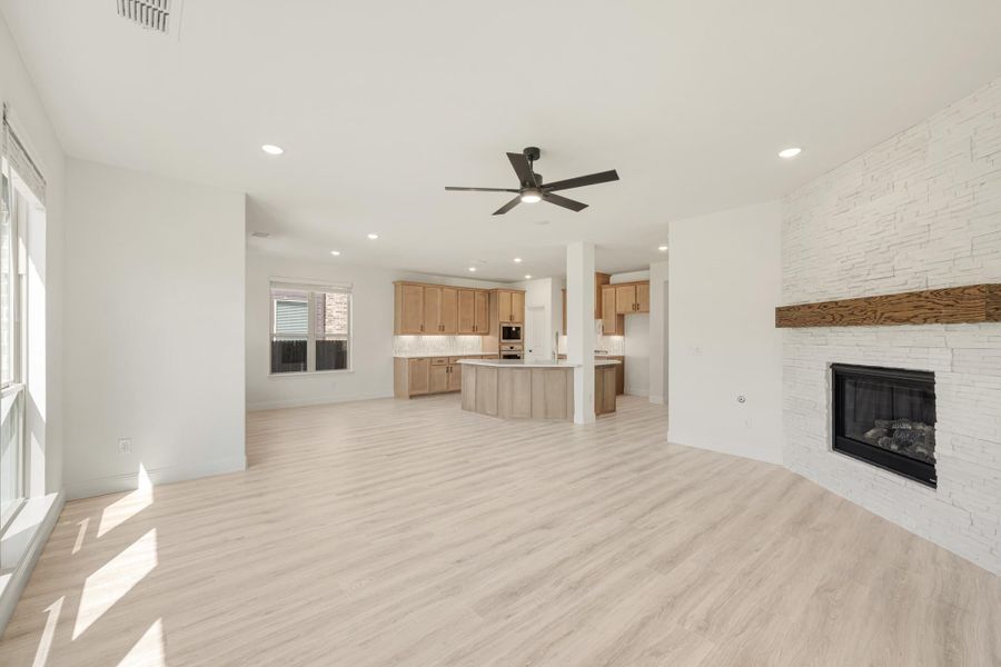 Open living area with stone fireplace, ceiling fan, and light wood floors open to kitchen with island