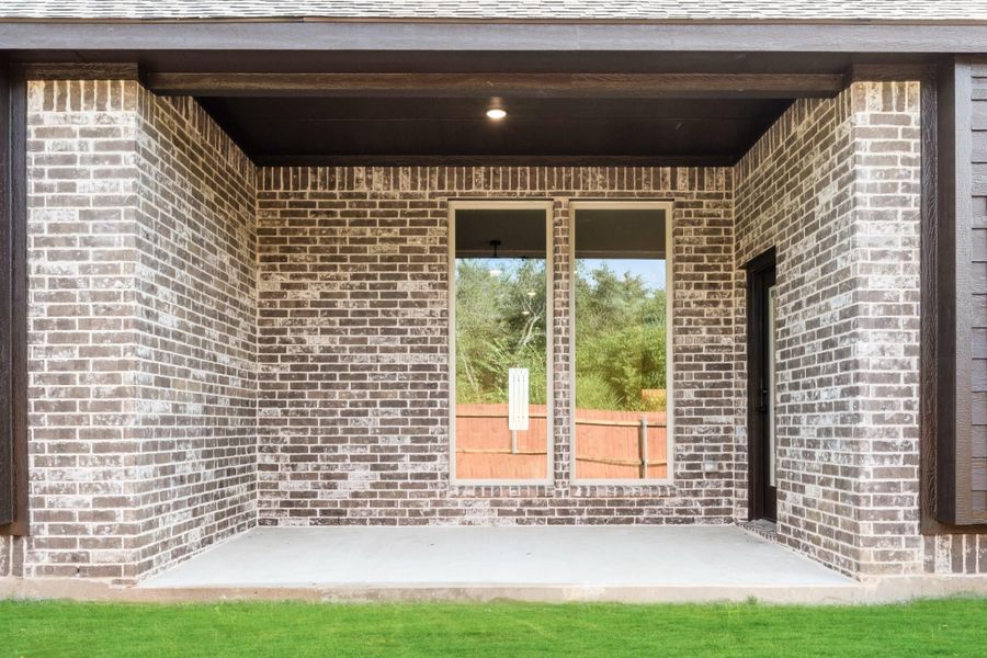 Covered back patio with brick walls, large window, black door, and concrete floor overlooking green lawn