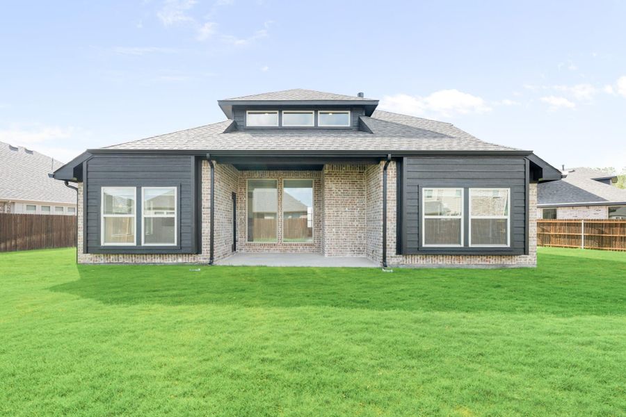 Rear view of single-story brick home with dark trim, covered patio, and large green backyard