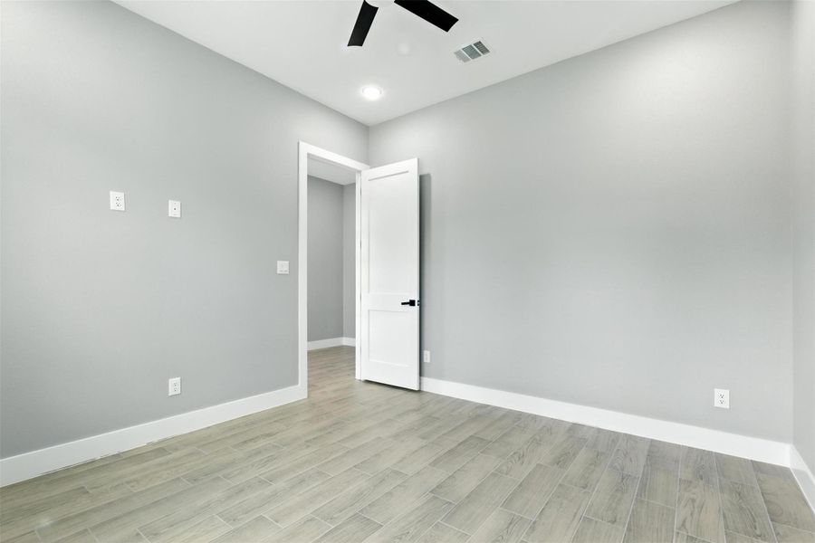 This photo showcases a bright, empty room featuring light gray walls, a white door, and light wood flooring. The room is equipped with ceiling lighting, a ceiling fan, and multiple outlets, offering a versatile space for customization.