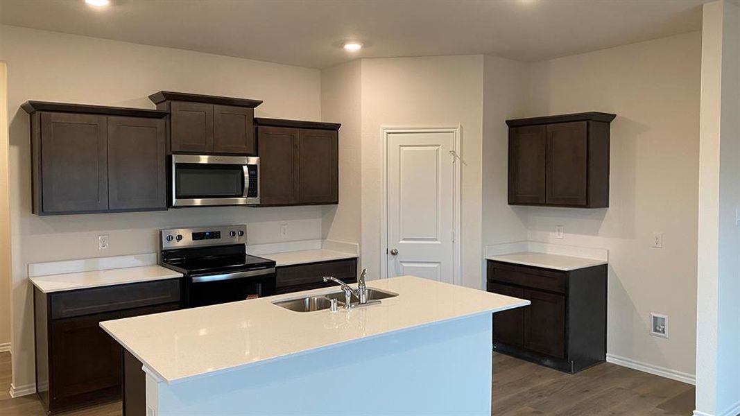 Kitchen featuring dark wood finish cabinets, stainless steel appliances, a kitchen island with sink, light stone counters, and dark wood-style flooring