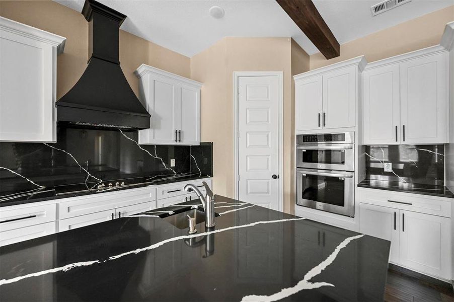 Kitchen with tasteful backsplash, white cabinets, stainless steel double oven, beam ceiling, and custom range hood