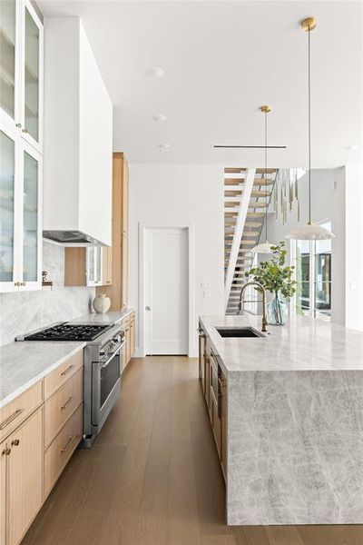 Modern kitchen featuring a large waterfall island with integrated sink, stainless steel range, light wood-finish cabinetry, white upper glass-front cabinets, and wood-finish flooring