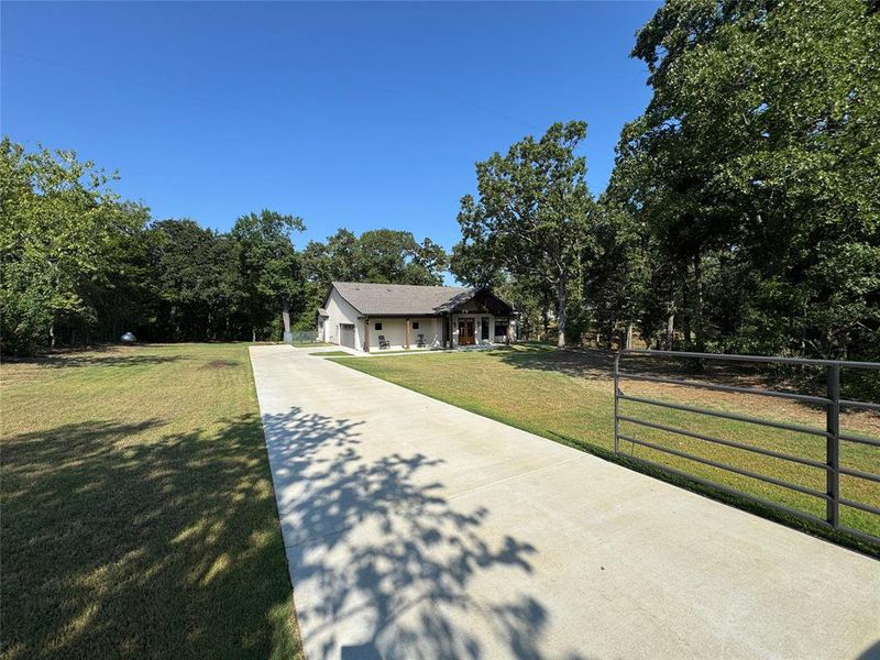 Single story home featuring concrete driveway, a front lawn, an attached garage, and view of wooded area Single story home featuring concrete driveway, a front lawn, an attached garage, and view of wooded area