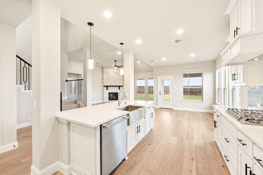 Open kitchen with white cabinets, center island, stainless appliances, and light wood floors opening to living area with stone fireplace.