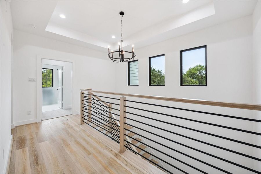 A light-filled upstairs landing showcases clean architectural lines with horizontal railing, wide-plank floors, and a trio of picture windows. A tray ceiling and modern chandelier add a touch of elegance to this airy transitional space. A light-filled upstairs landing showcases clean architectural lines with horizontal railing, wide-plank floors, and a trio of picture windows. A tray ceiling and modern chandelier add a touch of elegance to this airy transitional space.