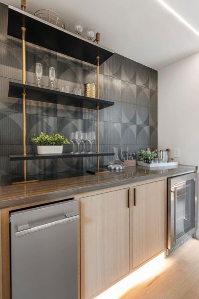 Thoughtfully designed wet bar featuring Eramosa Marble countertops and paired with a sleek backsplash adding texture to the space. Thoughtfully illuminated with warm overhead linear and under-shelf lighting. Thoughtfully designed wet bar featuring Eramosa Marble countertops and paired with a sleek backsplash adding texture to the space. Thoughtfully illuminated with warm overhead linear and under-shelf lighting.