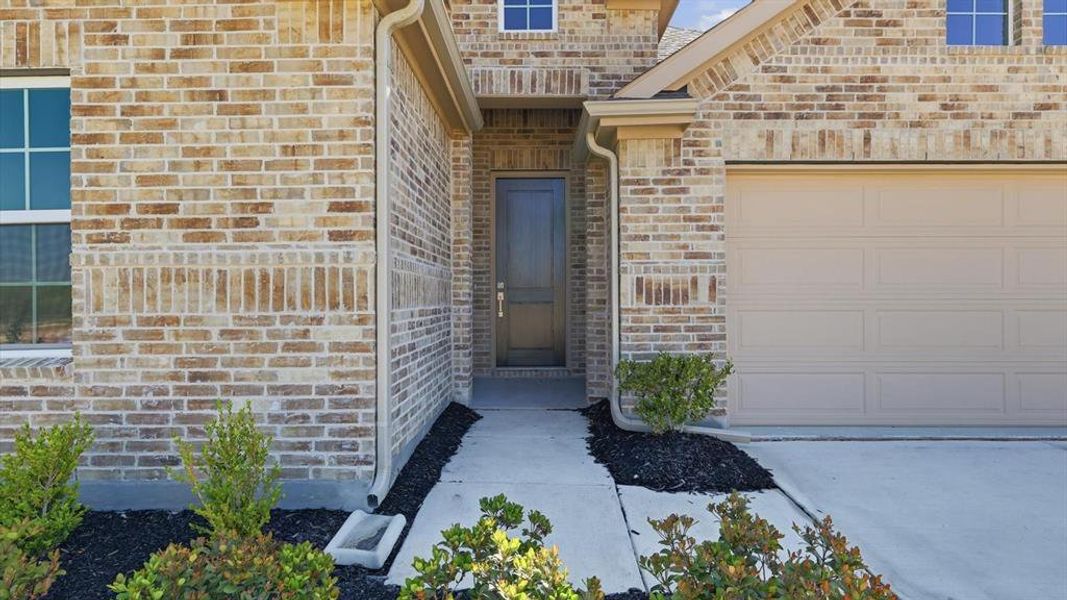 Brick exterior featuring a two-car garage, covered entry, front door, and mature landscaping