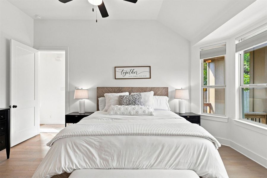 Hardwood floors and vaulted ceilings frame this bedroom illuminated by tall windows.