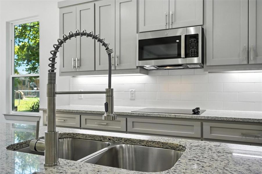 Kitchen featuring light stone countertops, stainless steel microwave, gray cabinetry, decorative backsplash, and black electric cooktop Kitchen featuring light stone countertops, stainless steel microwave, gray cabinetry, decorative backsplash, and black electric cooktop