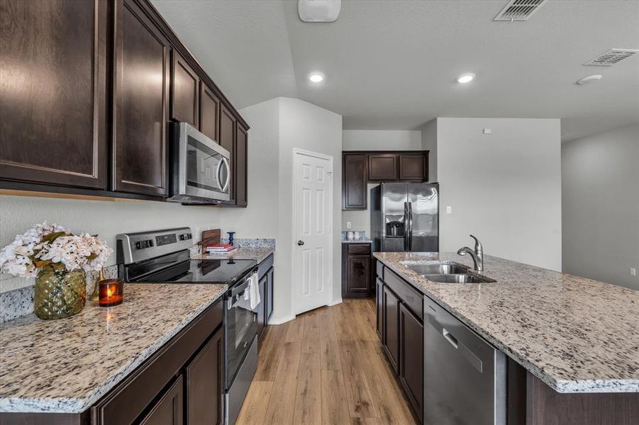 Kitchen featuring appliances with stainless steel finishes, dark brown cabinets, granite countertops, a kitchen island with sink, and generous corner pantry.