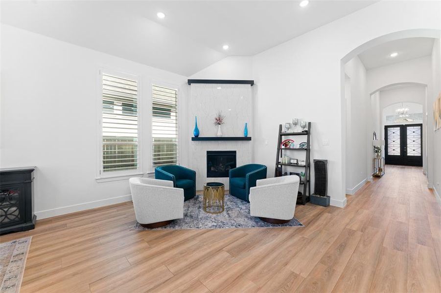 This living area features a fireplace with a white tiled surround and a dark wood mantel, wood plank flooring, and two windows with white louvered blinds