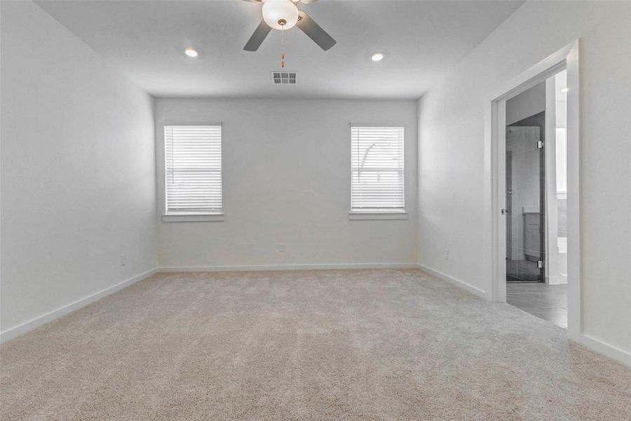 Spacious primary bedroom with soft carpet, neutral walls, recessed lighting, ceiling fan, and two windows for natural light. The private en-suite bath entrance adds convenience, while the open wall space and calm color palette make the room easy to personalize as a peaceful retreat.