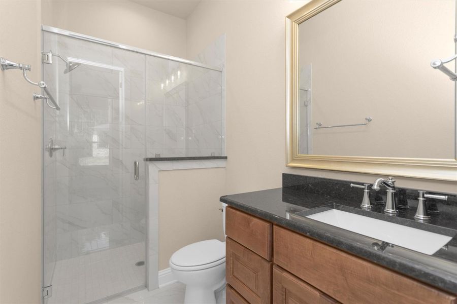 This sleek en-suite bathroom features a walk-in glass shower with polished tile surround, granite countertops, warm wood cabinetry, and a framed mirror—delivering both elegance and functionality.