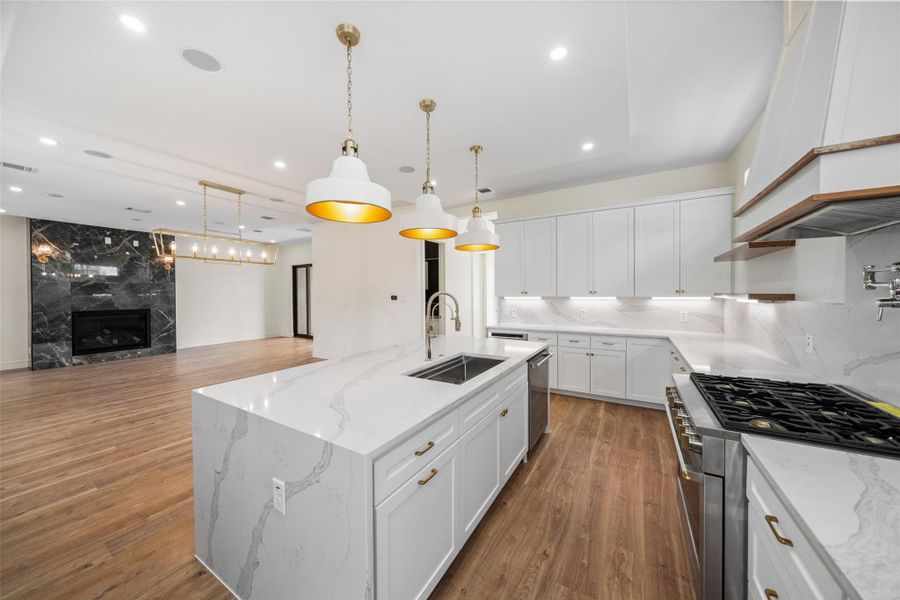 Kitchen features sleek white cabinetry, marble countertops, and a spacious island with a built-in sink. Elegant pendant lights hang above, and there's a high-end gas stove. The open layout flows seamlessly into a living area with a striking black marble fireplace. Wood flooring adds warmth and sophistication.