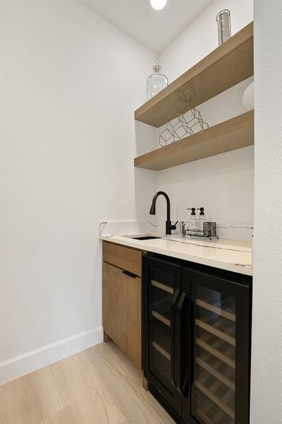 Indoor wet bar with wine cooler, open shelves, light wood-type flooring, light stone countertops, and brown cabinetry