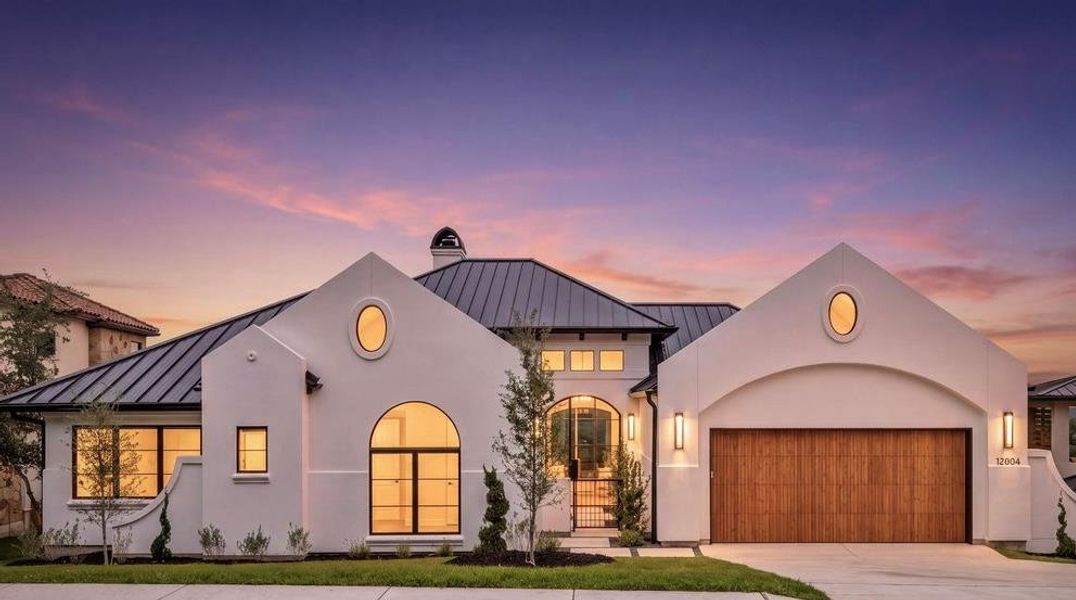 Contemporary stucco exterior with a dark metal standing seam roof, arched entryway, wood-finish garage door, and modern exterior lighting