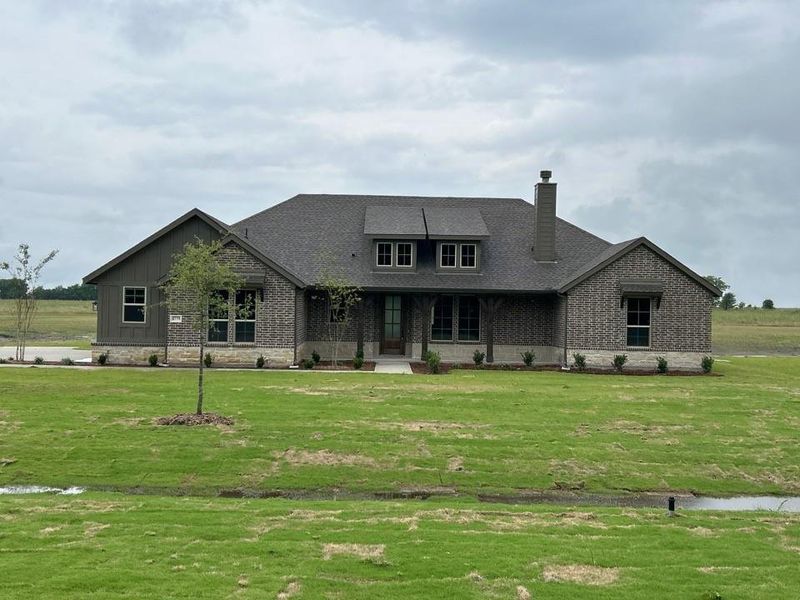 Single-story residence featuring a combination of gray brick and vertical board-and-batten siding, a dark shingle roof, and a stone water table Single-story residence featuring a combination of gray brick and vertical board-and-batten siding, a dark shingle roof, and a stone water table