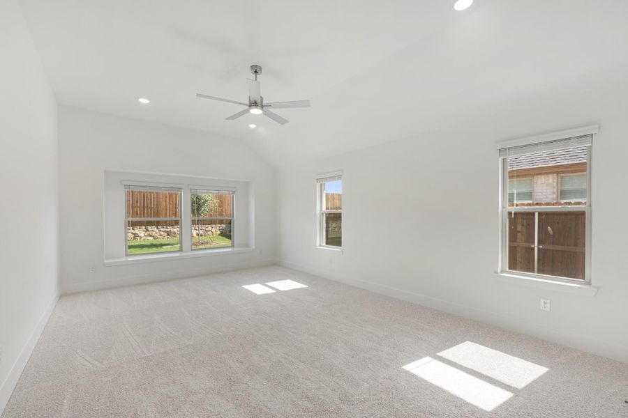 Empty bedroom with vaulted ceiling, ceiling fan, carpet flooring, and three windows with backyard views Empty bedroom with vaulted ceiling, ceiling fan, carpet flooring, and three windows with backyard views