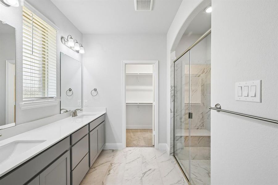 Bathroom featuring a double vanity with gray cabinetry, a walk-in shower with clear glass enclosure and stone-look tile, and a spacious walk-in closet Bathroom featuring a double vanity with gray cabinetry, a walk-in shower with clear glass enclosure and stone-look tile, and a spacious walk-in closet