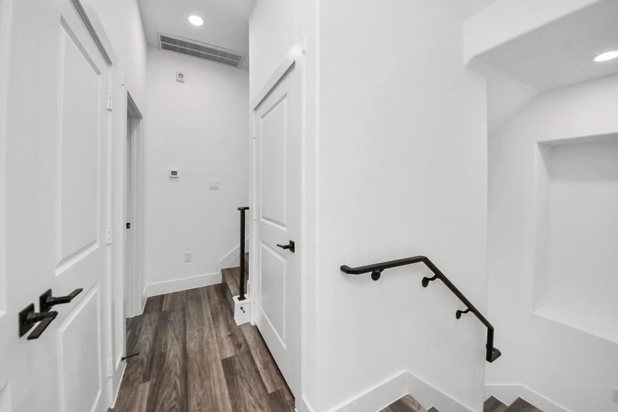 This photo shows a modern, well-lit hallway featuring white walls, vinyl flooring, and a staircase with a black handrail. There are multiple doors and recessed lighting, creating a clean, contemporary look. This photo shows a modern, well-lit hallway featuring white walls, vinyl flooring, and a staircase with a black handrail. There are multiple doors and recessed lighting, creating a clean, contemporary look.