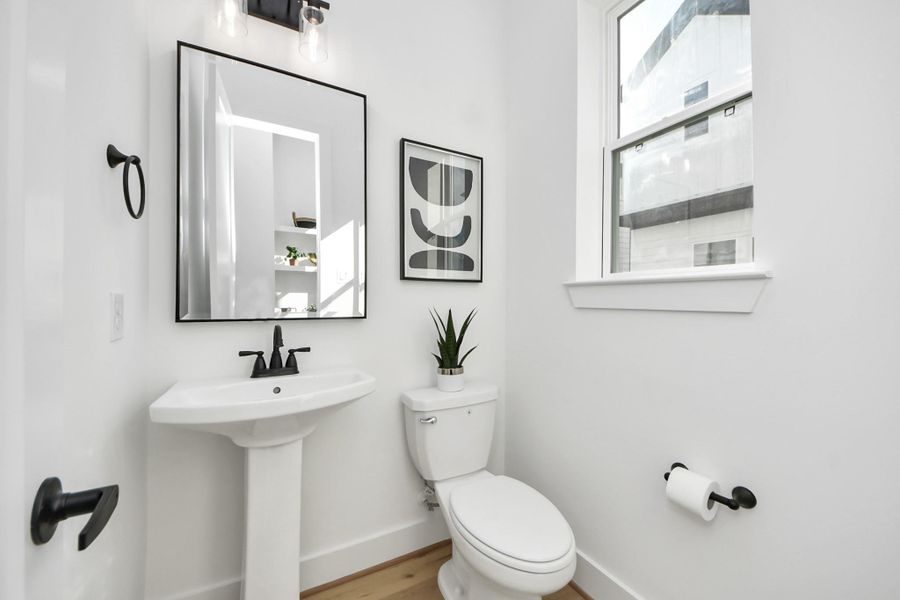 This is a modern, bright bathroom with a minimalist design. It features a pedestal sink with a black faucet, a wall-mounted mirror, and a toilet. The room is accented with a small plant, geometric artwork, and natural light from a window. This is a modern, bright bathroom with a minimalist design. It features a pedestal sink with a black faucet, a wall-mounted mirror, and a toilet. The room is accented with a small plant, geometric artwork, and natural light from a window.