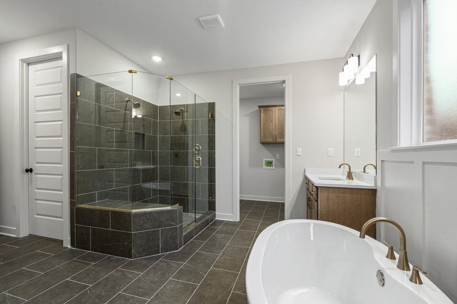 Spacious bathroom featuring a freestanding soaking tub with brass fixtures, a glass-enclosed shower with dark tile surround and built-in bench, dual vanities with white countertops and brass faucets, and grey tiled flooring Spacious bathroom featuring a freestanding soaking tub with brass fixtures, a glass-enclosed shower with dark tile surround and built-in bench, dual vanities with white countertops and brass faucets, and grey tiled flooring