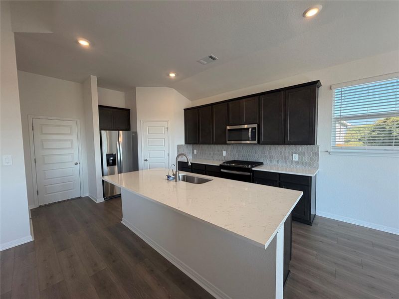Kitchen with appliances with stainless steel finishes, tasteful backsplash, light stone counters, a kitchen island with sink, and dark wood-type flooring