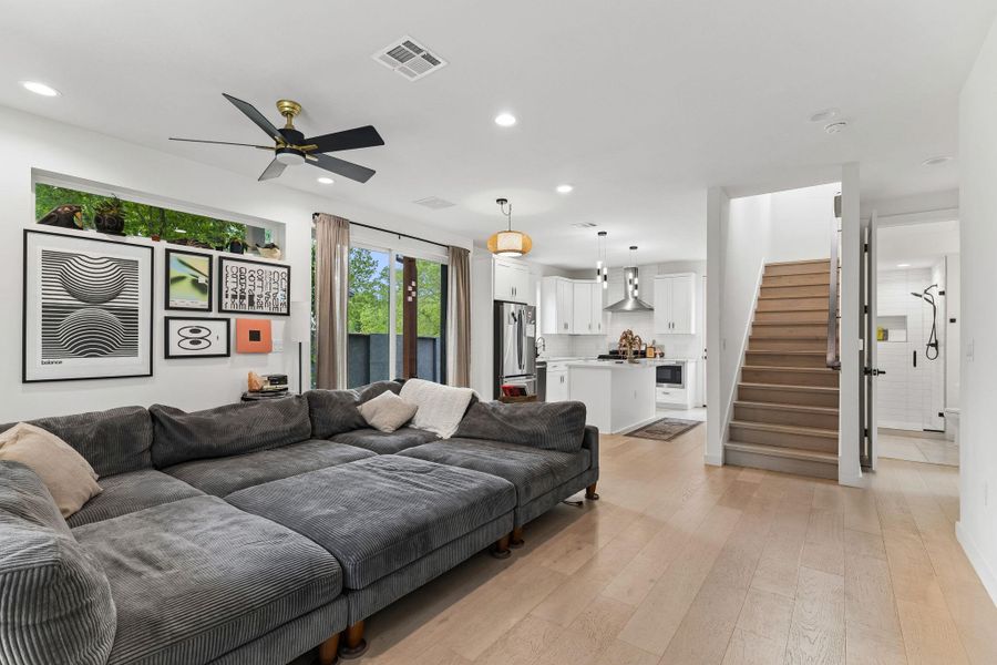 An expansive view of the open-concept main floor. The living area is anchored by a large, comfortable sectional sofa and features light wood-look flooring and a modern black-and-gold ceiling fan. The room flows seamlessly into the kitchen and toward the staircase leading to the upper levels.