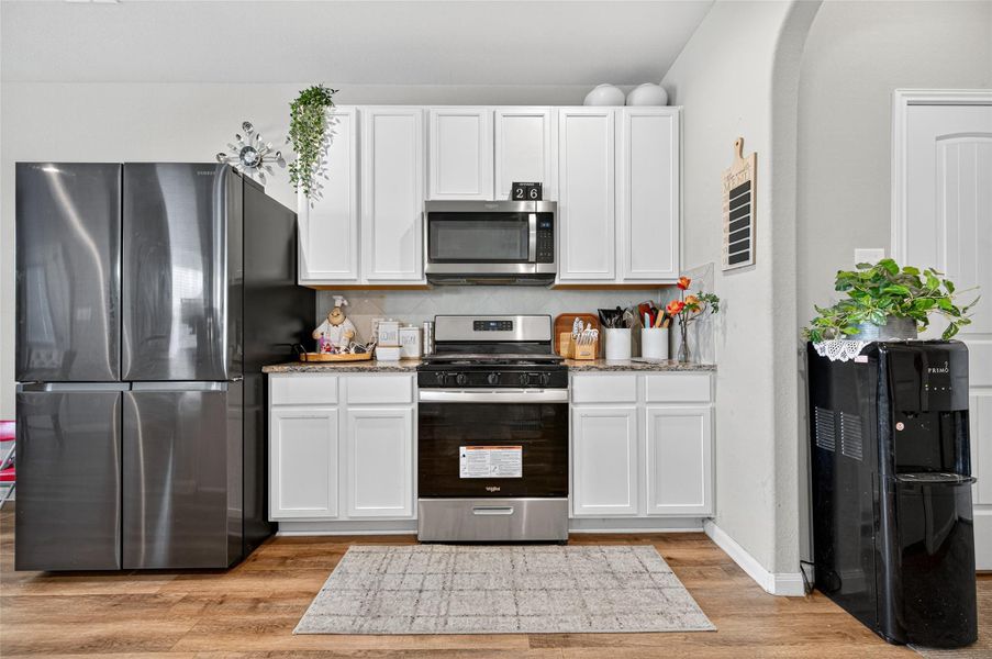 Modern kitchen with stainless steel appliances, white cabinetry, and wood flooring. The space includes a sleek refrigerator, gas stove, microwave, and ample countertop space, accented with plants and decor for a welcoming feel.