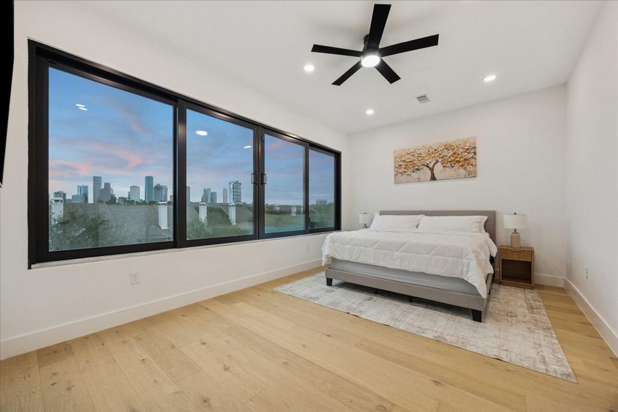 Spacious primary suite featuring wide-plank wood flooring and expansive windows showcasing stunning Houston skyline views.