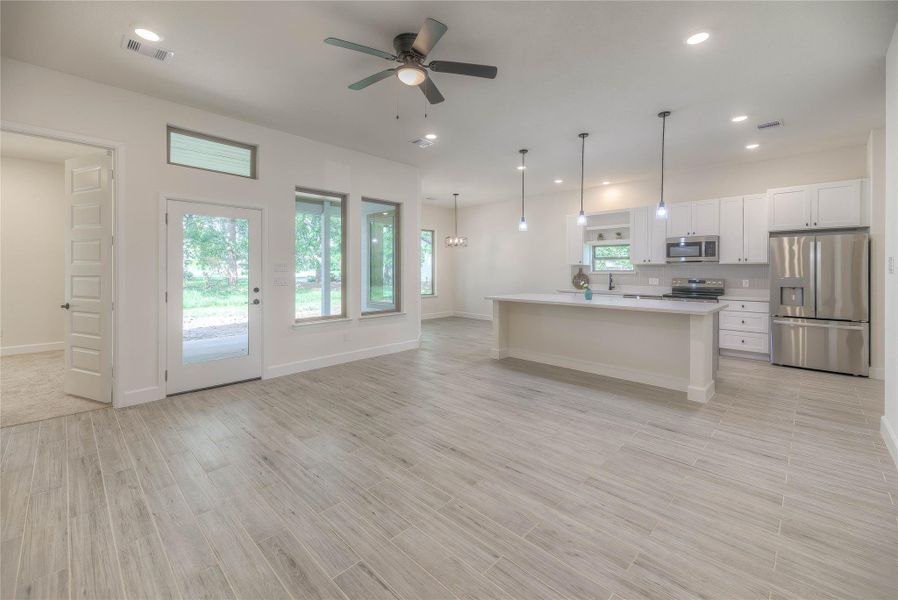 This spacious open-concept living area features modern tile flooring, a ceiling fan, and ample natural light from large windows. The kitchen boasts stainless steel appliances, pendant lighting, and a large island with plenty of counter space.